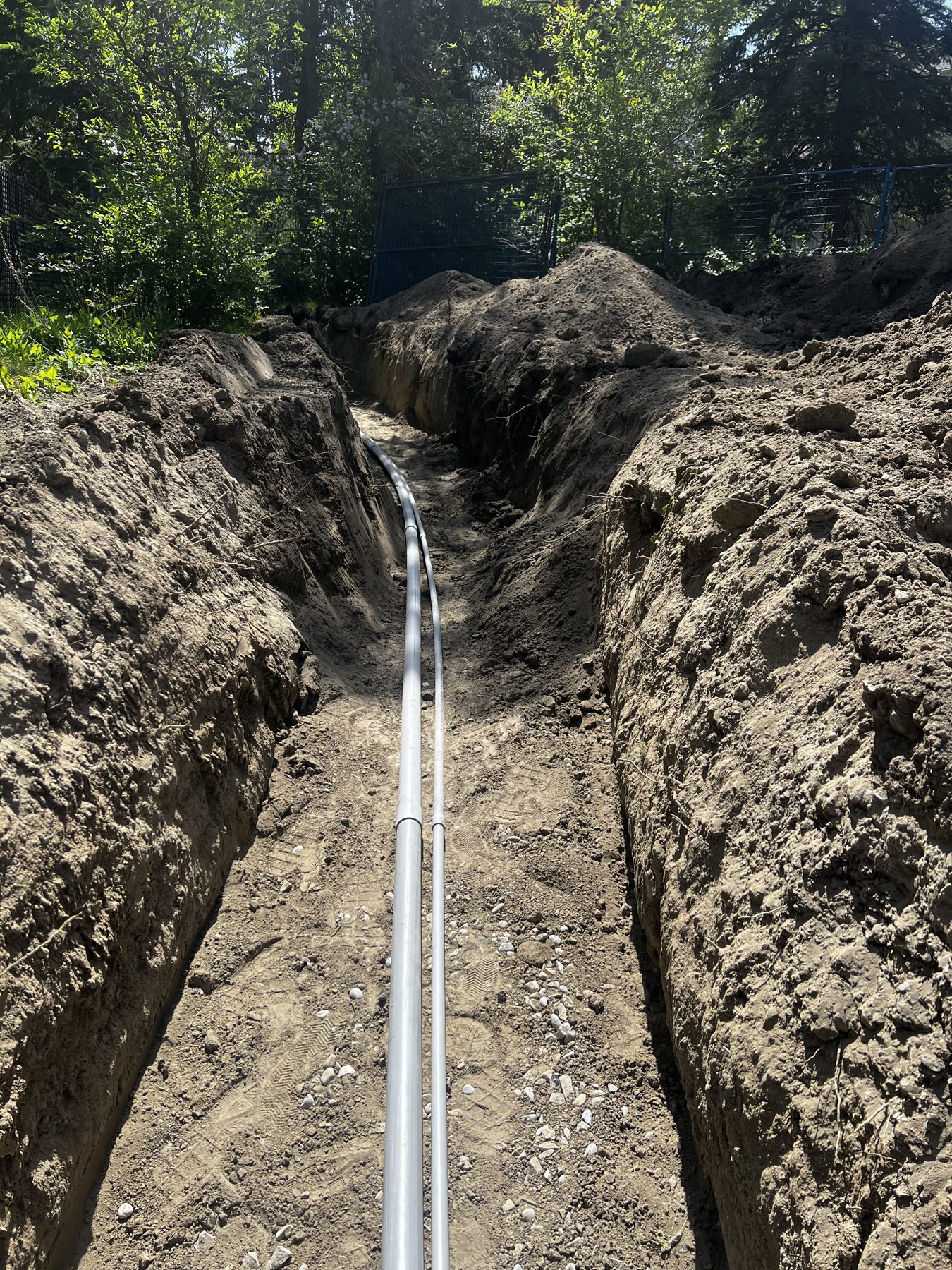 Trench dug with pipes installed, dirt mounds, trees and fence visible — 18 Varsplain Place Northwest by Onsite Contracting Services
