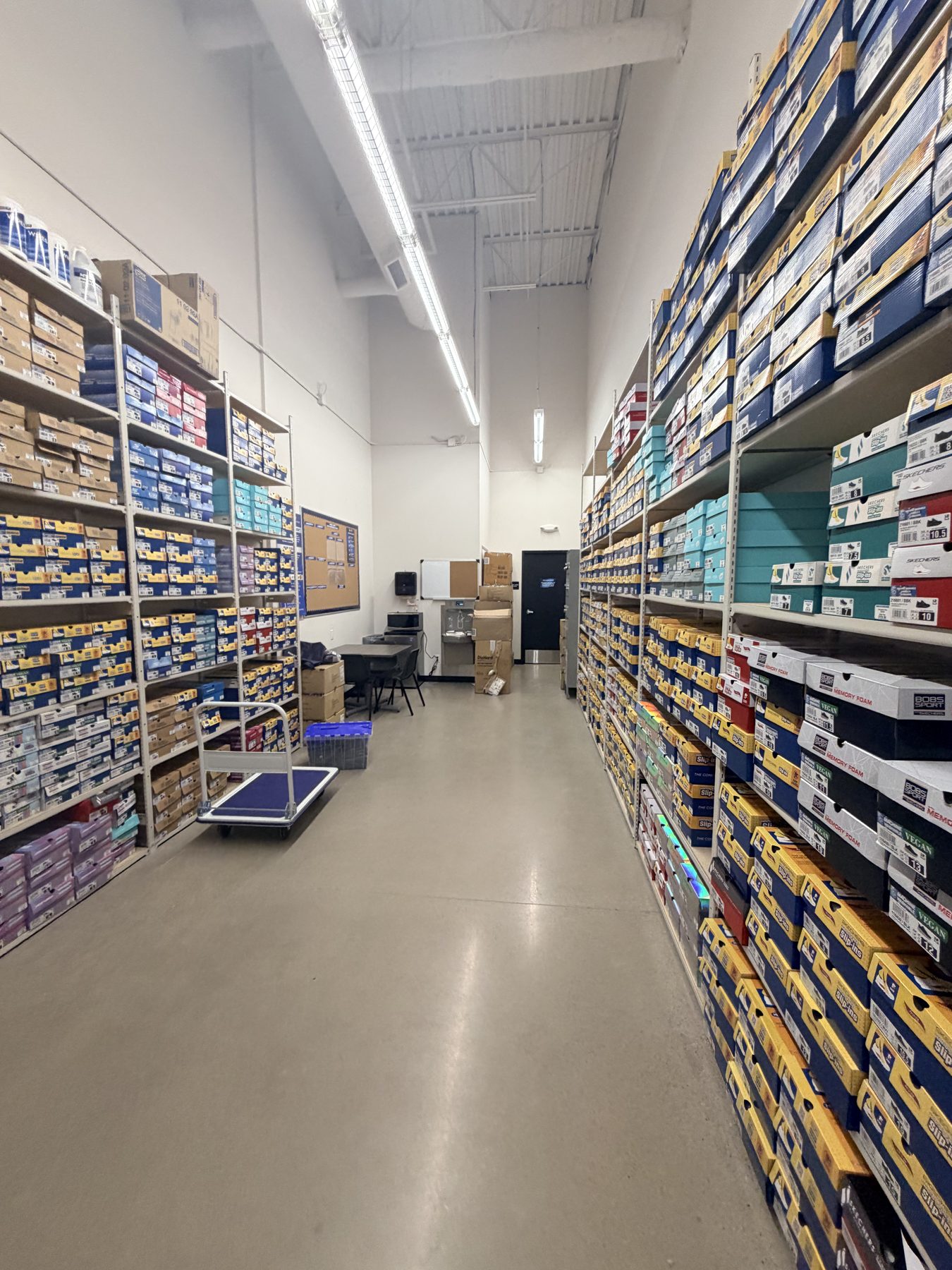 Store aisle stocked with shoe boxes on shelves and a blue cart — Skechers (Signal Hill)