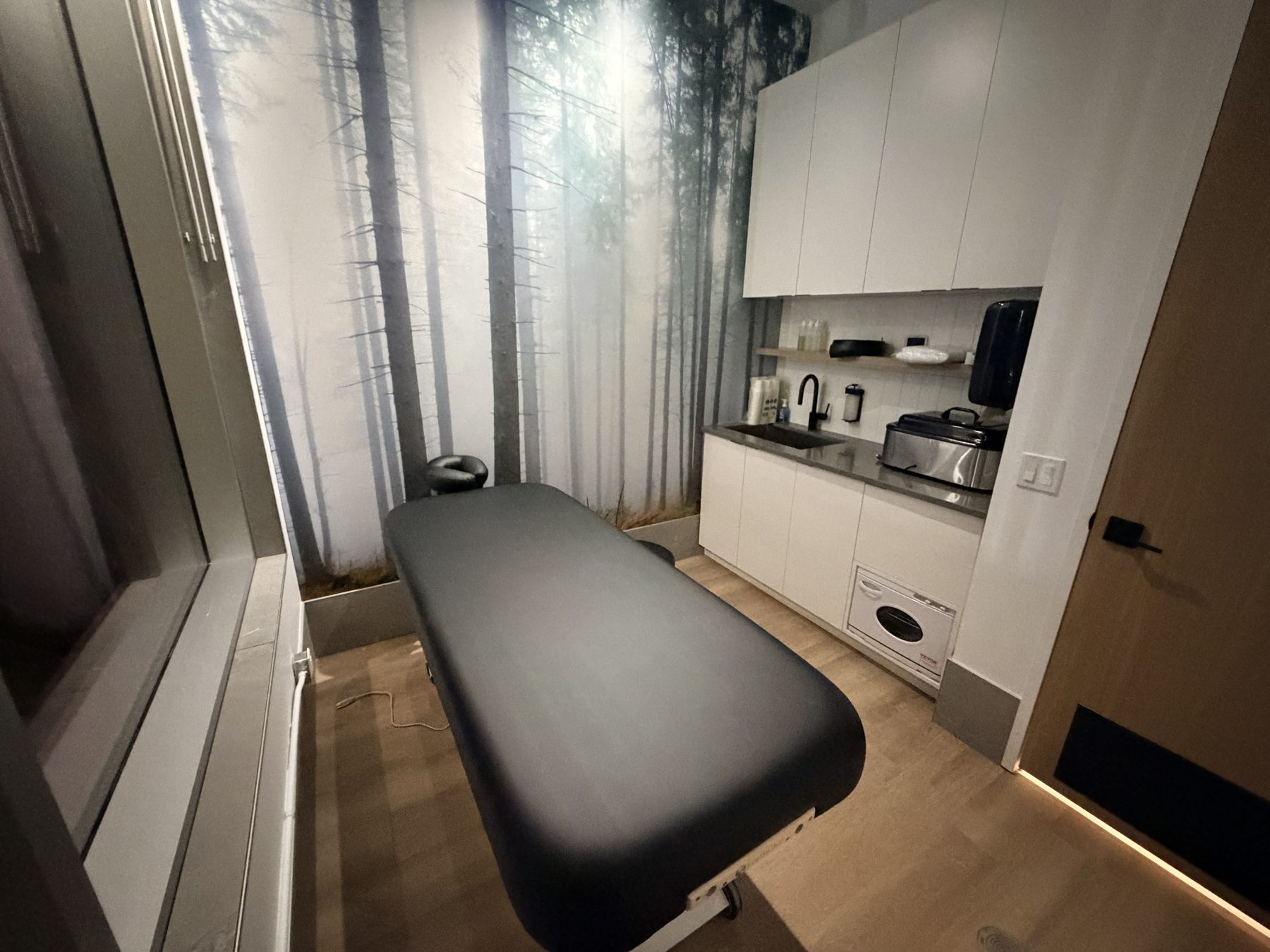 Massage room with forest wallpaper, black massage table, and sink counter — Cedar Steam and Spa (Trinity Hills)