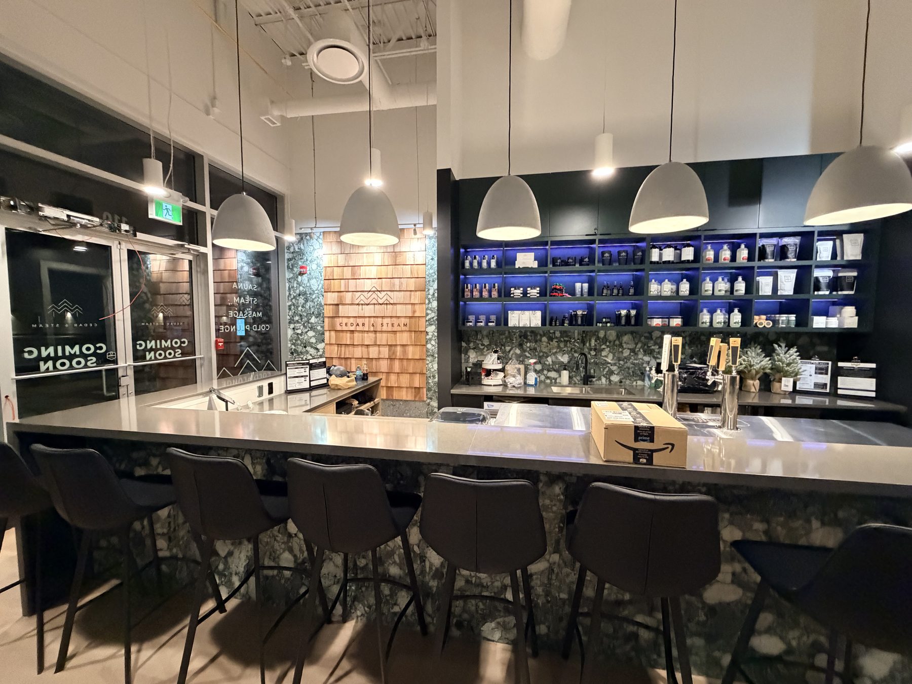 Bar with grey counter, marble base, and black stools, shelves stocked — Cedar Steam and Spa (Trinity Hills)