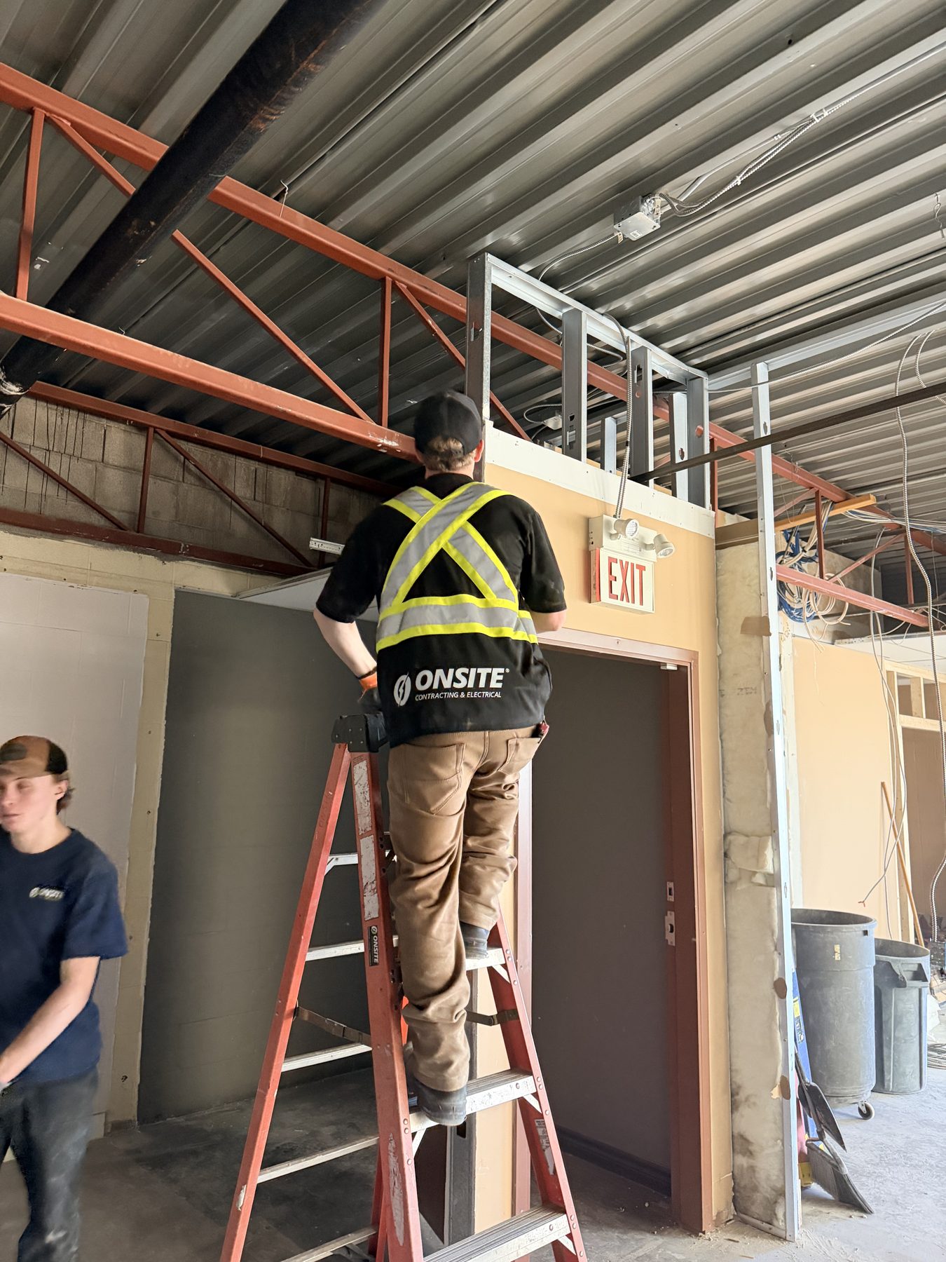 Worker on ladder installs metal framing near exit sign and exposed ceiling joists — The Barron Venue