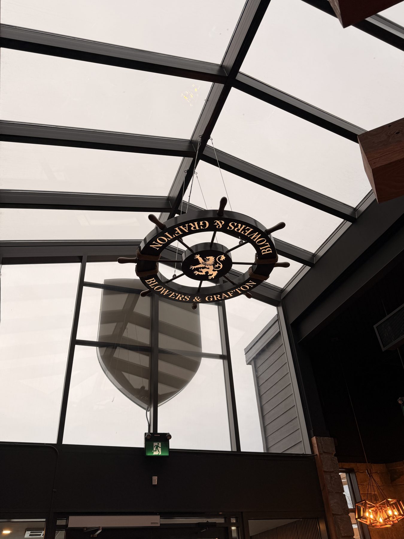Ship's wheel sign hangs from glass ceiling, partially obscured shield artwork — Blowers and Grafton (Red Deer)