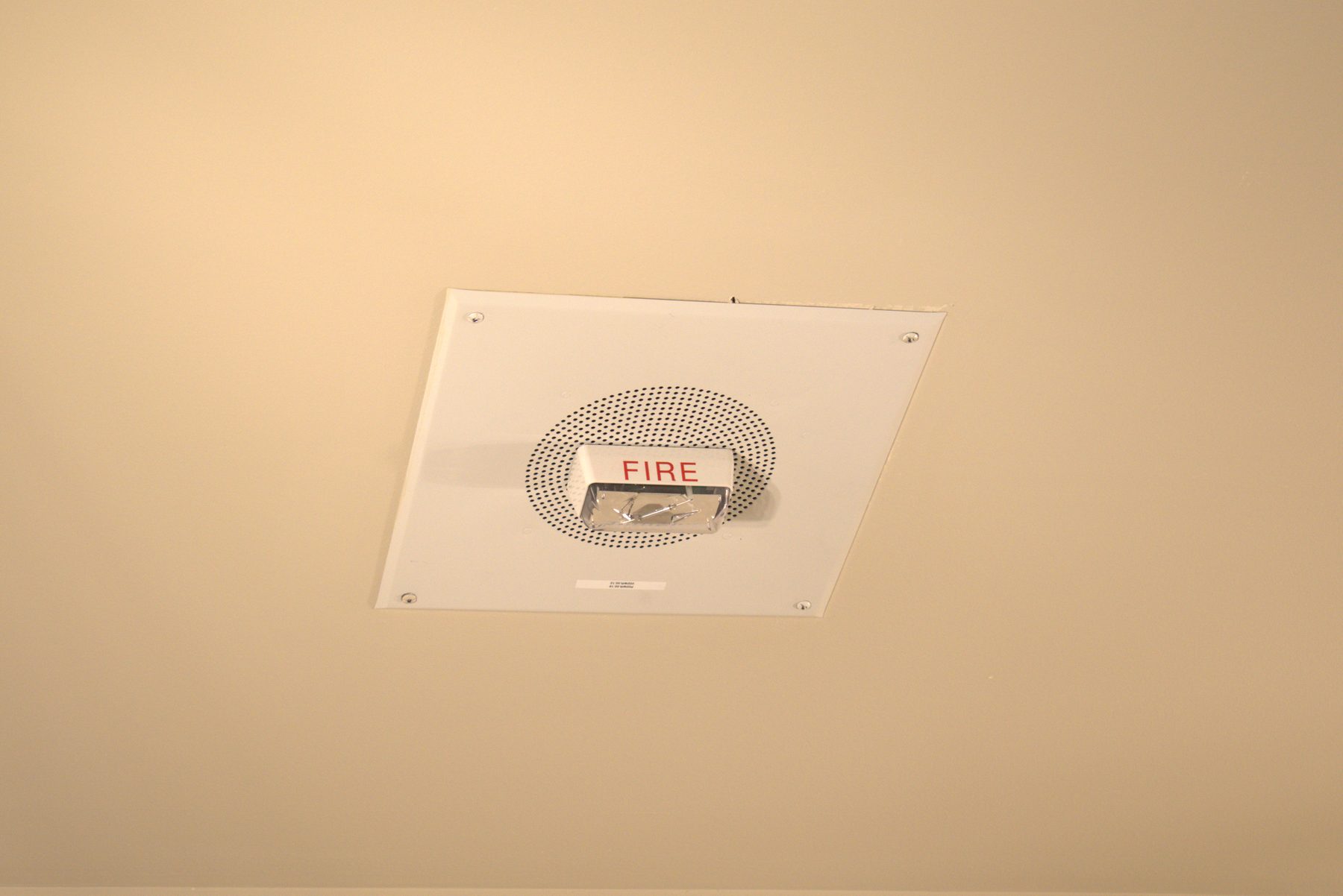 Newly installed white fire alarm speaker and strobe on a beige ceiling — Leela Spa (Bankers Hall)