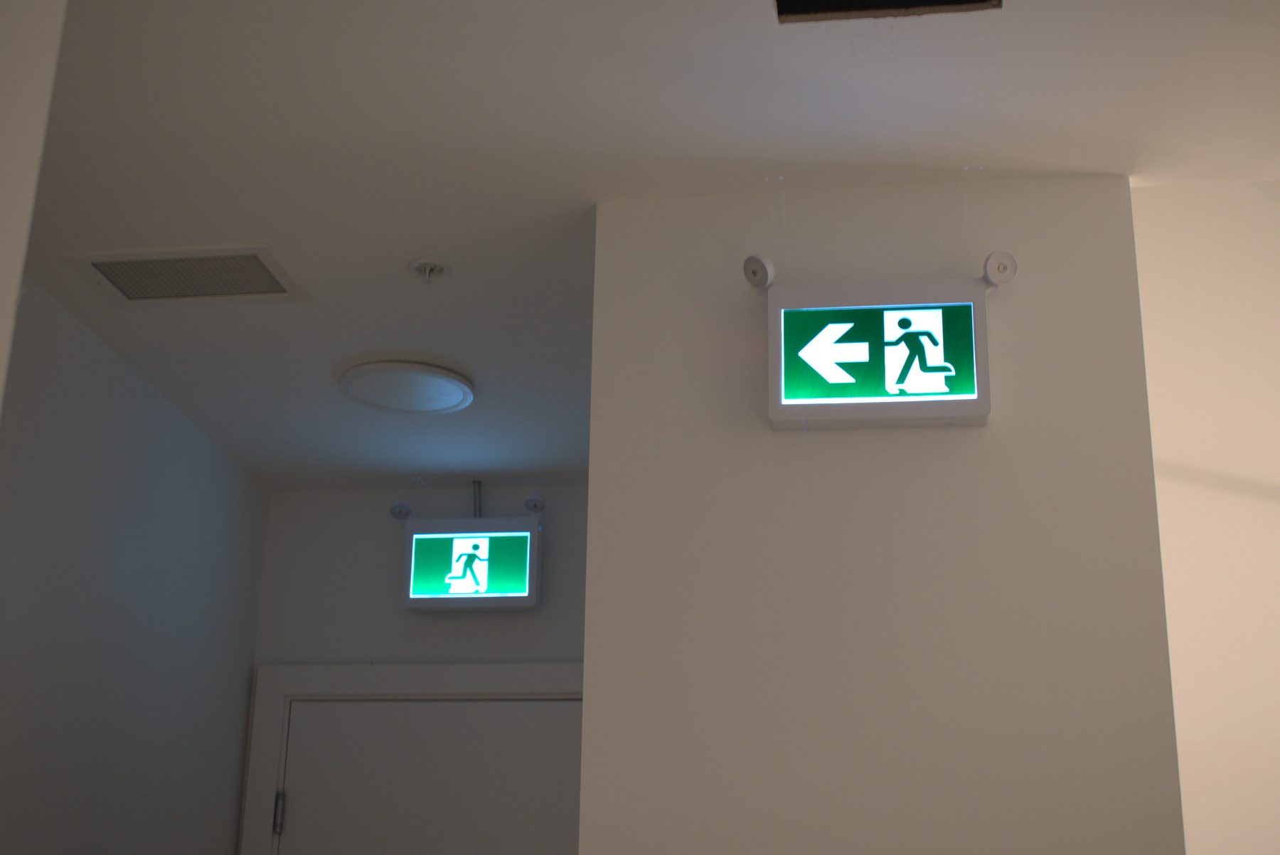 Two illuminated green exit signs installed on white walls, one large, one small — Big Buns (Bowness)