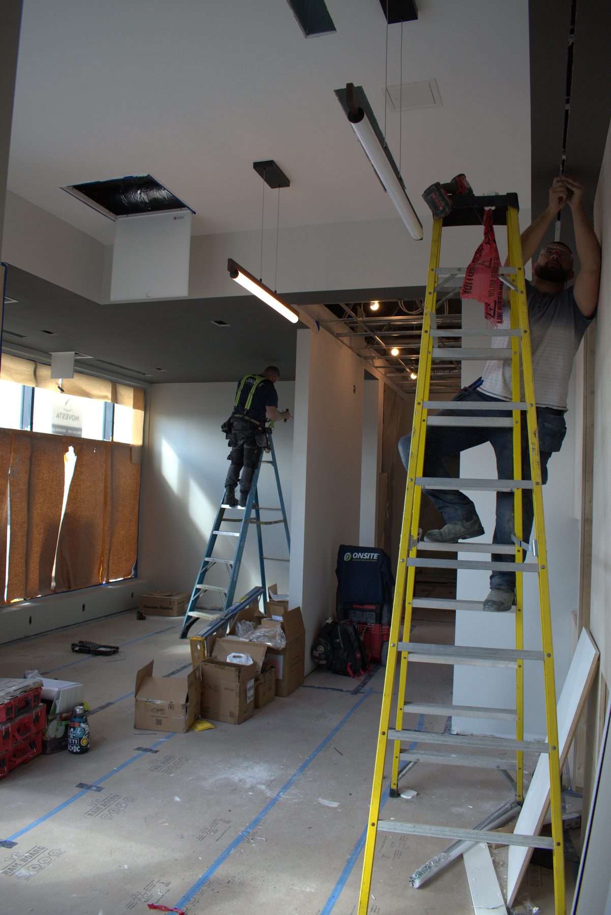 Workers on ladders install ceiling lights and trim in unfinished room — ONE8T (West District)