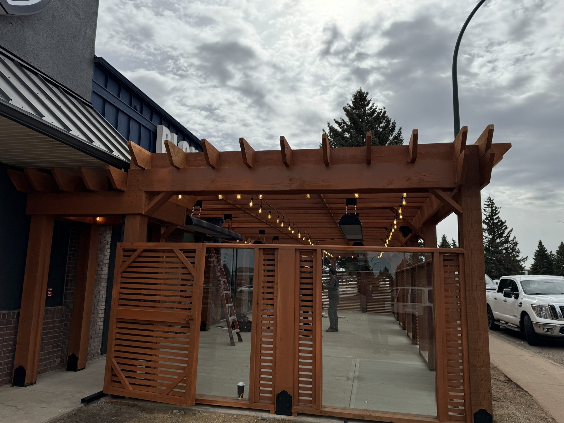 Wooden pergola with string lights and glass panels under construction — Blowers and Grafton (Medicine Hat)