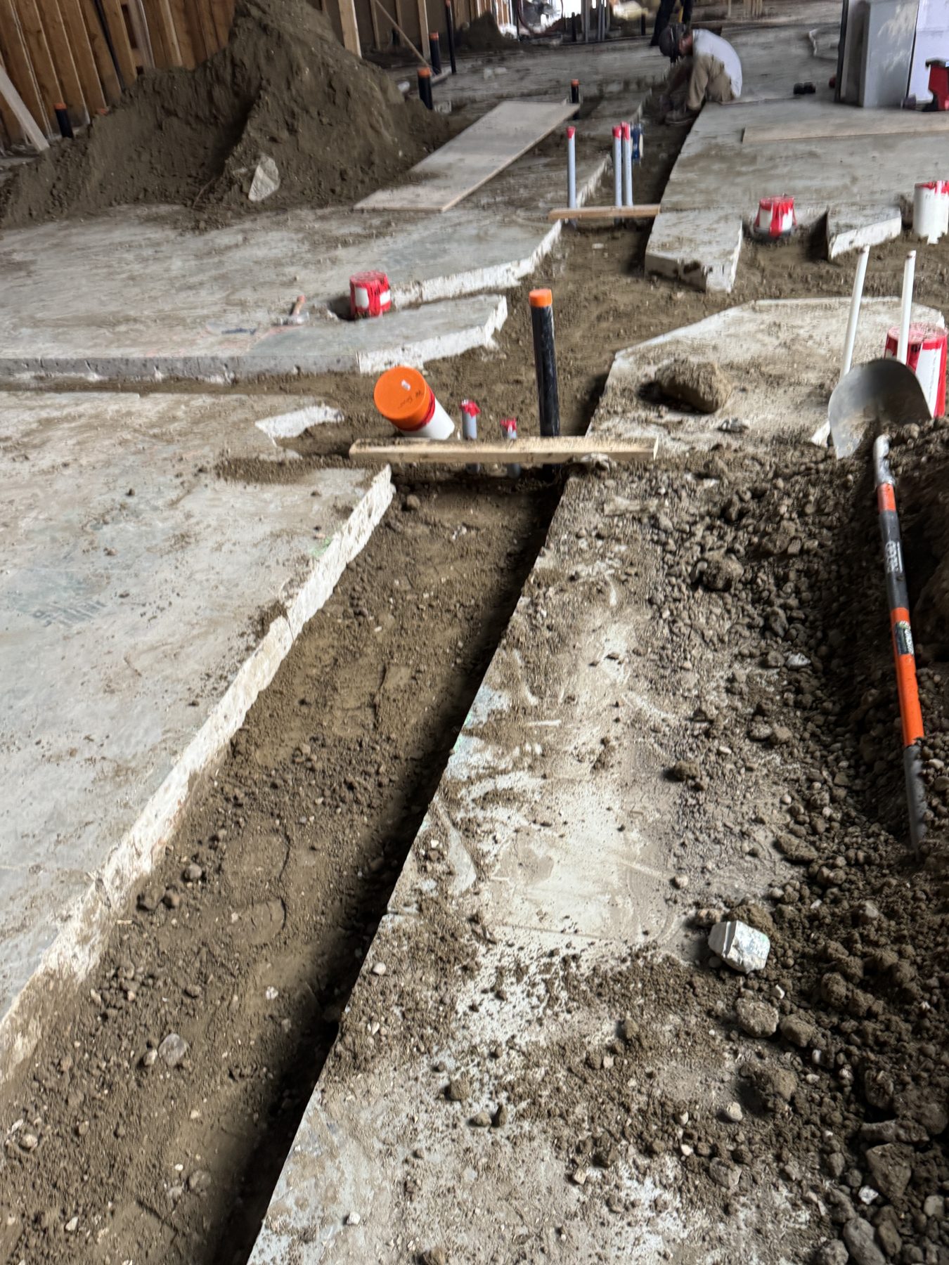 Rough plumbing trenches cut into concrete floor, pipes and dirt visible — Blowers and Grafton (Red Deer)