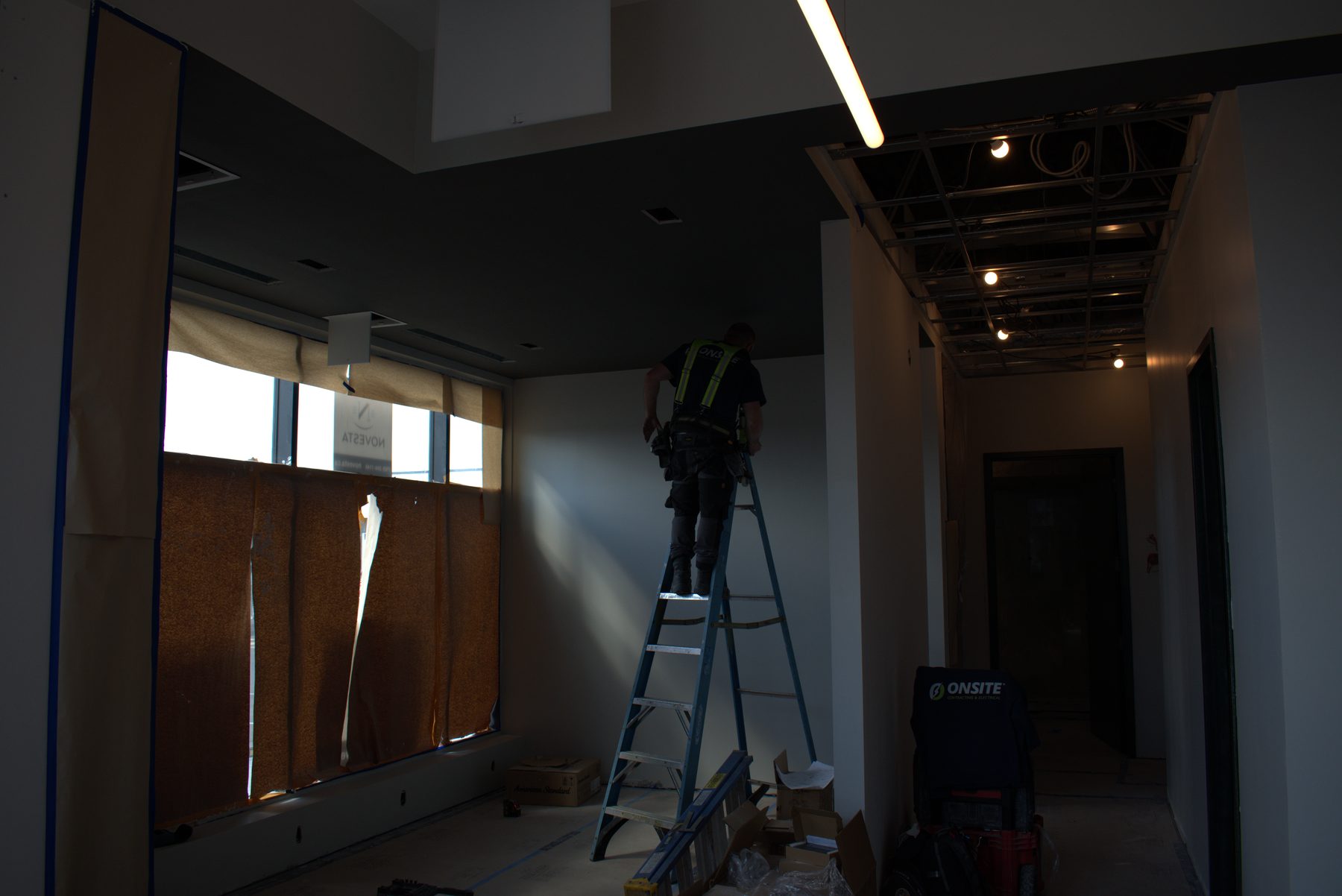 Construction worker on ladder, bare ceiling grid, drywall, and unfinished interior — ONE8T (West District)